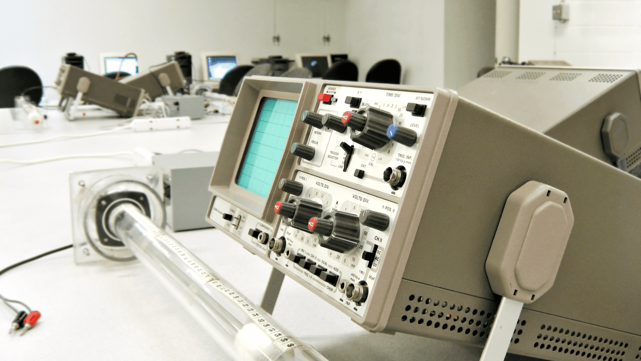 Analogue oscilloscope with time-base and volts-per-division controls on a lab bench beside a graduated glass tube